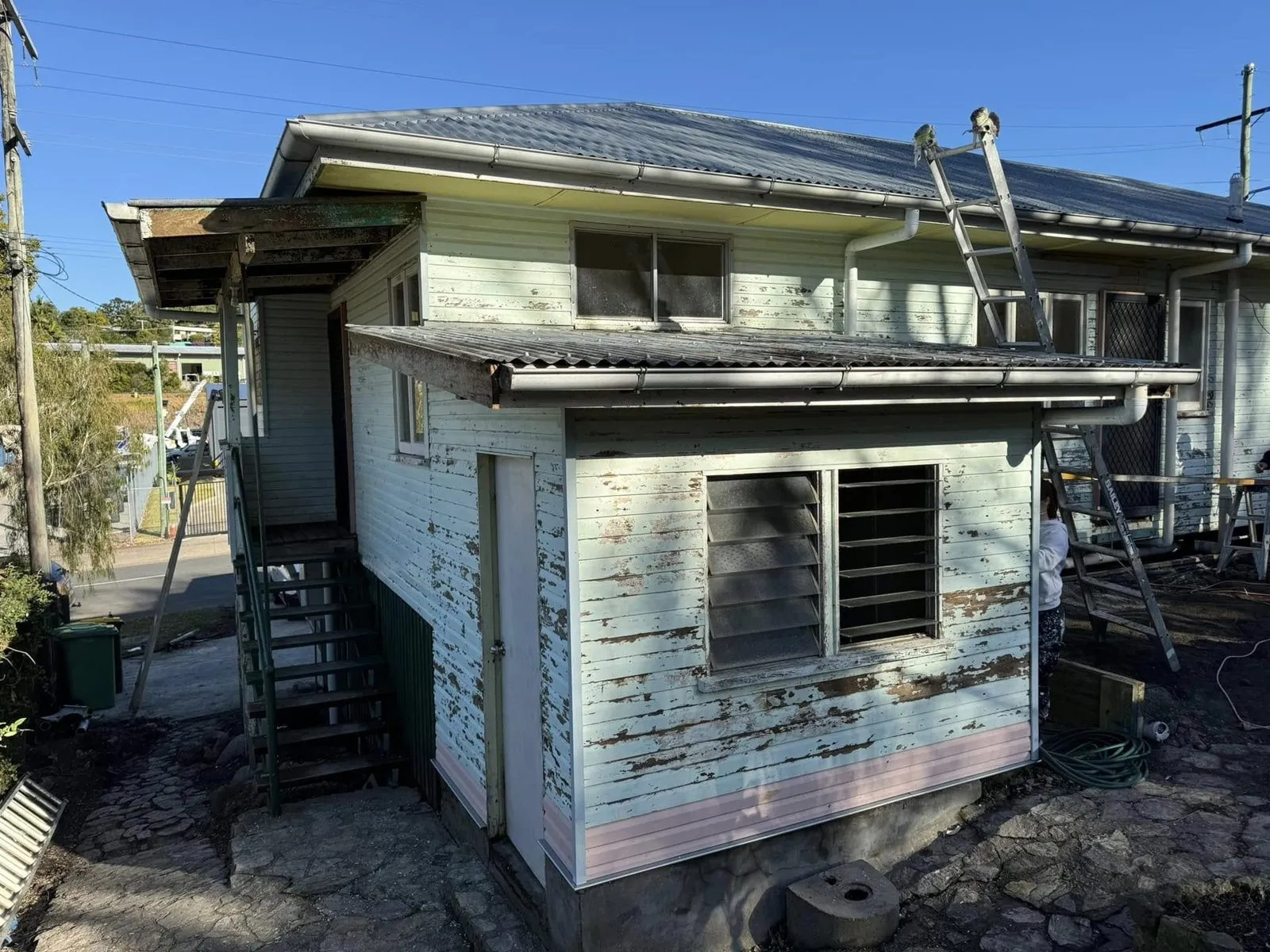 Weatherboard house before painting, with badly flaking pale paint and bare timber showing on the walls, and ladders set up nearby
