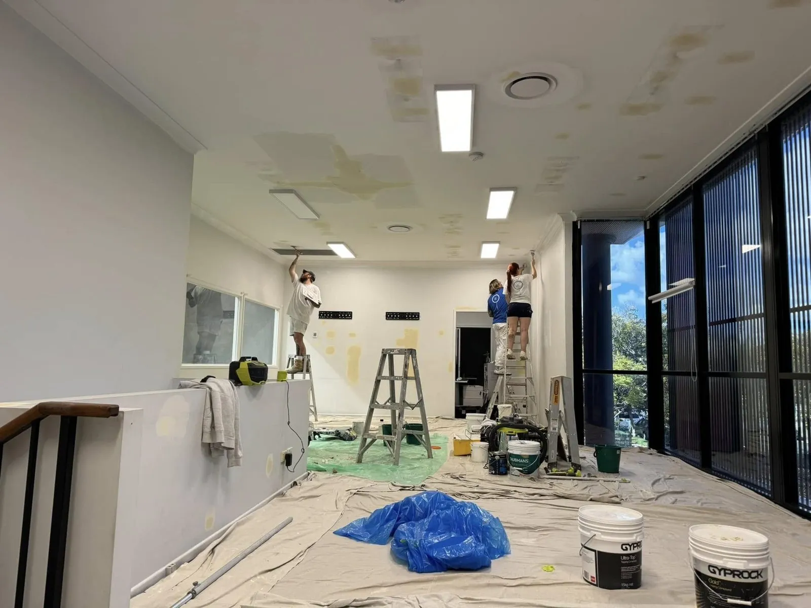Painters on ladders patching stained, water-damaged ceiling plaster in an office with drop sheets on the floor