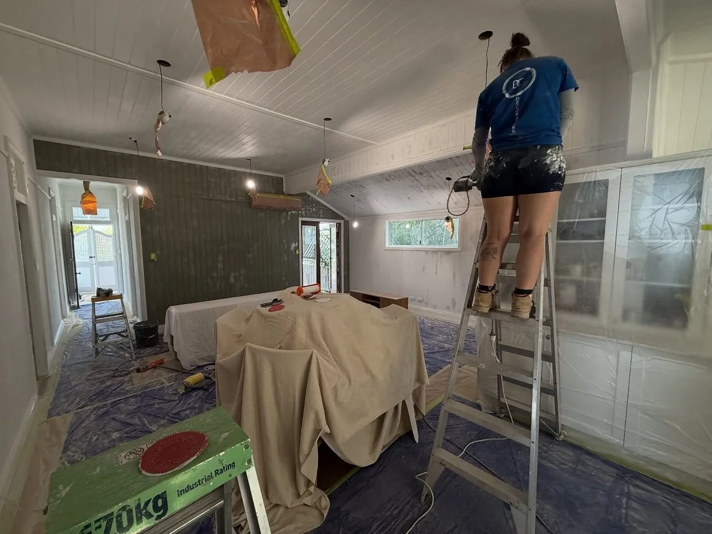 DC painter on a ladder sanding a lined ceiling, with the furniture and floor covered below