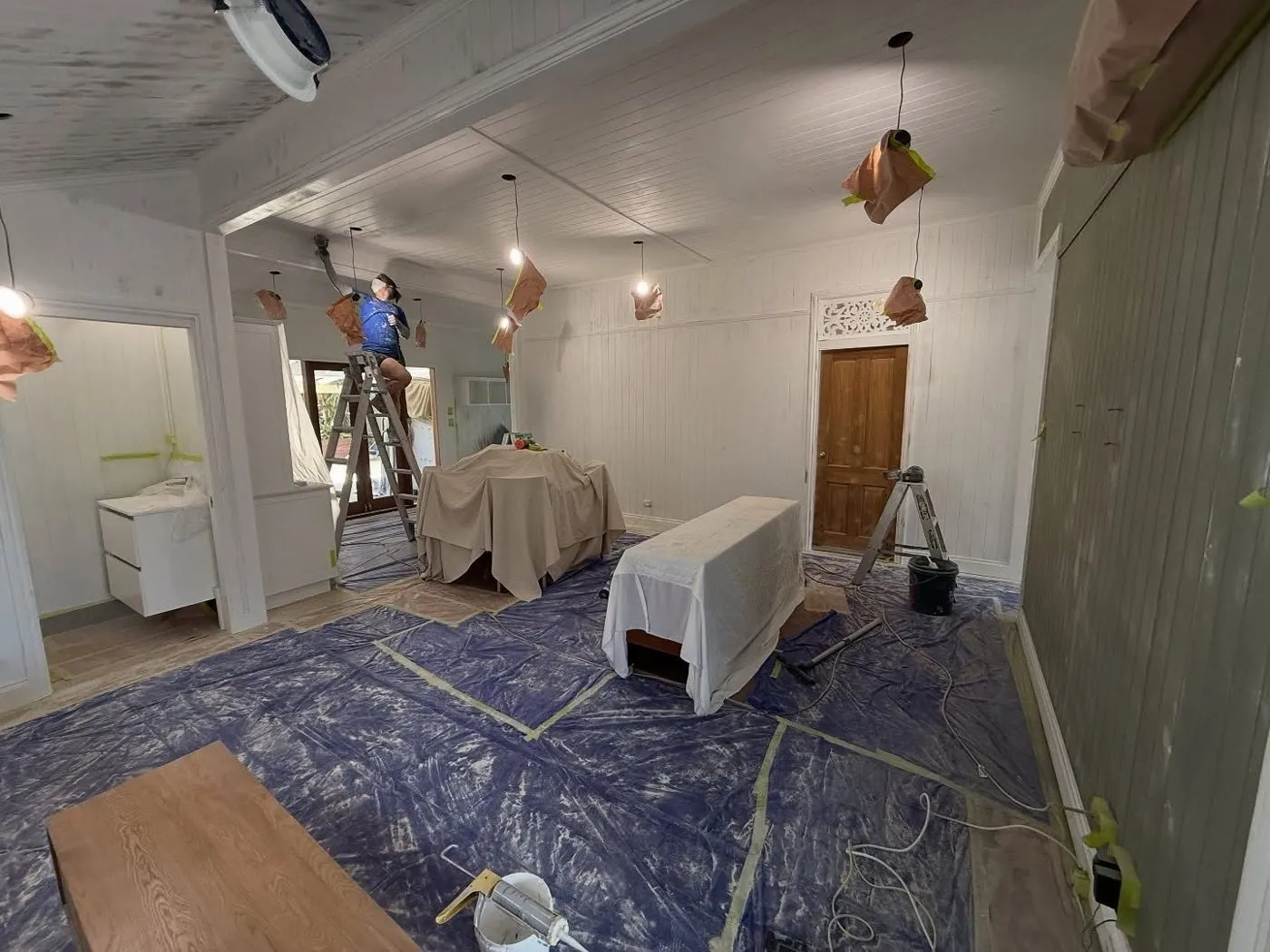 Room with the floor covered in sheeting, furniture under drop sheets and a painter working on the ceiling